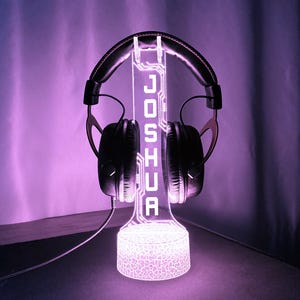 May include: A glowing purple headphone stand with the name "JOSHUA" illuminated in white. The stand holds a pair of black headphones. The base has a textured pattern, and the overall design is modern and tech-inspired.