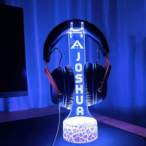 May include: A white LED light-up headphone stand with a custom name "JOSHUA" and a triangle logo. The stand has a textured base and is lit up in blue.