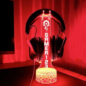 以下が含まれることがあります： 赤いLEDライトが、白いロゴと「GAMER123」の文字が書かれた黒いゲーミングヘッドセットスタンドを照らしています。スタンドはテクスチャ加工されたベースを持っています。