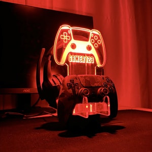 Peut inclure: Un support de jeu rouge éclairé avec un support pour manette et casque. Le support présente une manette de jeu translucide avec le texte "GAMER123".