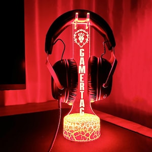 Op de afbeelding: Een zwart-wit gamertag-bord met een leeuwenlogo, verlicht in rood en geel, met een zwarte koptelefoon die op de standaard rust.