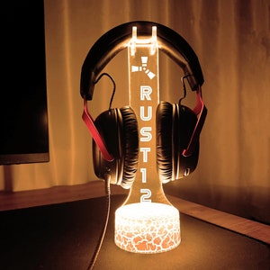May include: A black and red gaming headset sits on a white acrylic stand with the word "RUST12" printed on it. The stand has a white light emanating from the base.