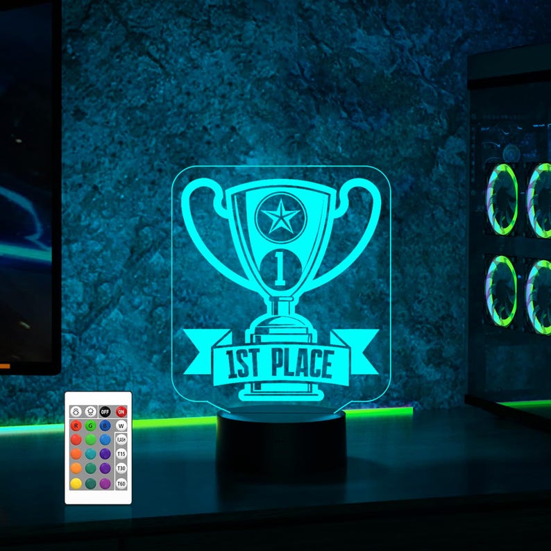 Puede incluir: Una luz nocturna impresa en 3D azul y blanca con forma de trofeo con el texto "1st Place" en una cinta. La luz nocturna est&aacute; sobre una base negra y tiene un mando a distancia con m&uacute;ltiples opciones de color.