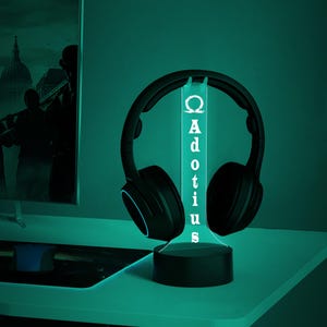May include: Black headphones resting on a glowing LED headphone stand. The stand is black with the word "Adotius" vertically displayed in white. The stand is on a white surface, with a computer monitor in the background.