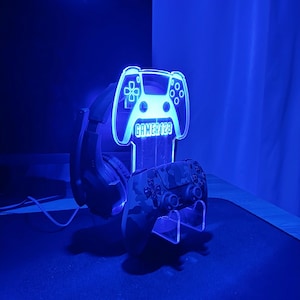Op de afbeelding: Een blauw verlichte gaming headset stand in de vorm van een controller. De stand toont de tekst "GAMER123" en ondersteunt een camouflage-patroon controller en een zwarte headset. De opstelling staat op een donkere ondergrond.