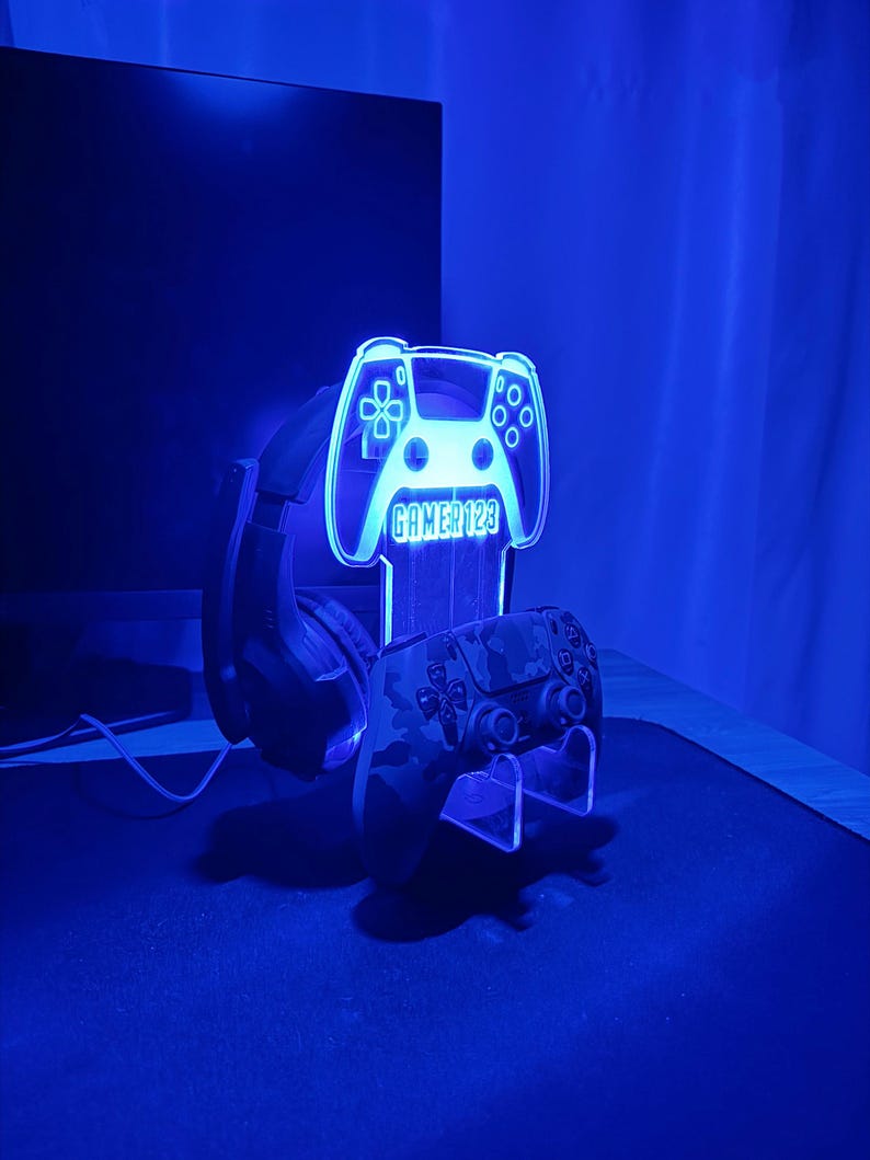 Op de afbeelding: Een blauw verlichte gaming headset stand in de vorm van een controller. De stand toont de tekst "GAMER123" en ondersteunt een camouflage-patroon controller en een zwarte headset. De opstelling staat op een donkere ondergrond.