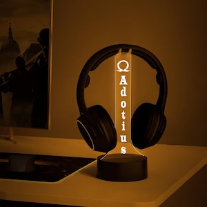 May include: A black headset rests on a custom-made headphone stand. The stand is illuminated with a warm glow, featuring the name "Adotius" in white lettering. The stand is on a white surface.