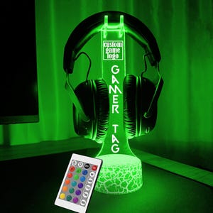 May include: A green LED light stand with a cracked white base. The stand holds a pair of black headphones. The stand has a white rectangular sign with the text "custom game logo GAMER TAG". The stand comes with a remote control with multiple color options.