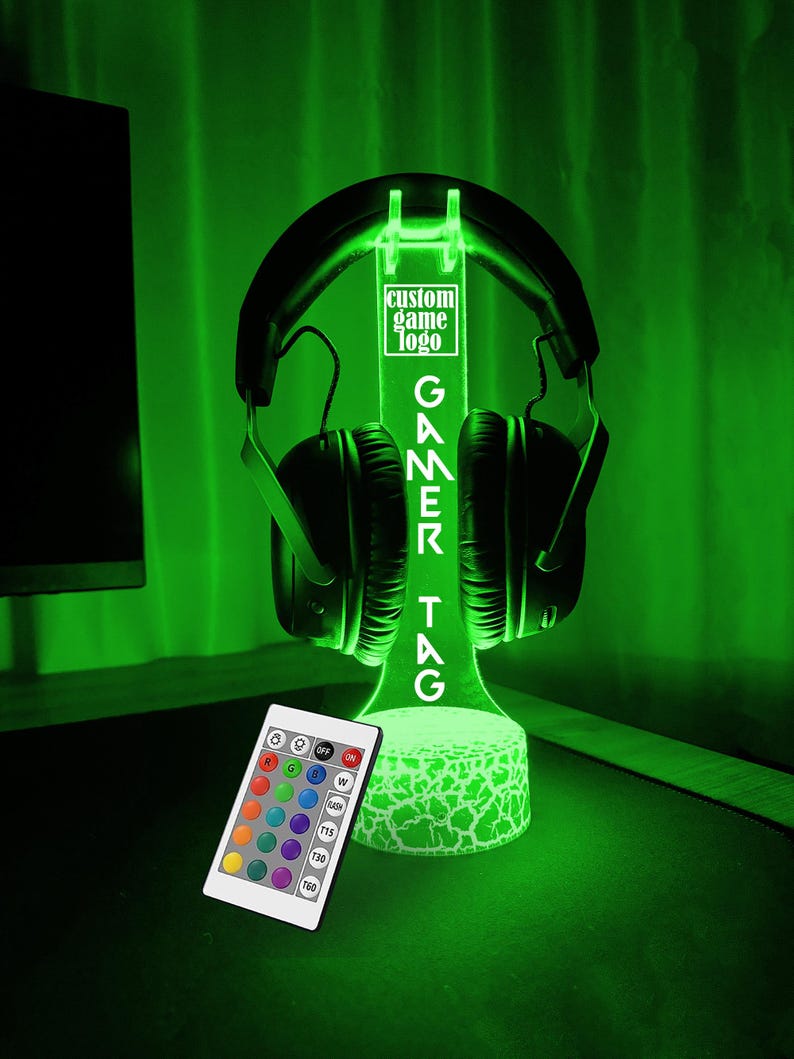 May include: A green LED light stand with a cracked white base. The stand holds a pair of black headphones. The stand has a white rectangular sign with the text "custom game logo GAMER TAG". The stand comes with a remote control with multiple color options.