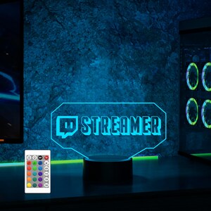 Op de afbeelding: Een blauw neon-bord met de tekst "STREAMER" en een Twitch-logo. Het bord staat op een zwarte basis en heeft een afstandsbediening met gekleurde knoppen.