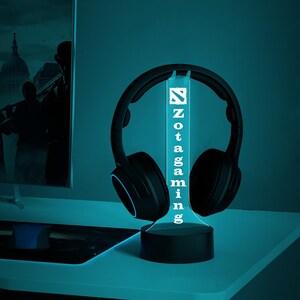 May include: A black gaming headset with a blue light on the earcups is displayed on a black stand with a white Zotac Gaming logo. The stand is on a blue surface.
