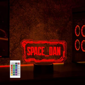 May include: A red LED light with a black base, displaying the text "SPACE_DAN" in a futuristic font. The light is controlled by a remote with multiple color options.