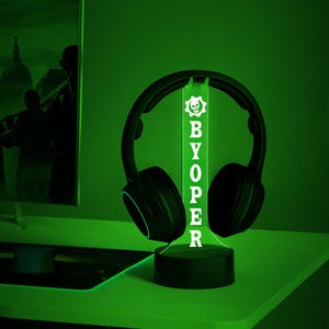 Op de afbeelding: Een groen LED-licht met een schedel en gekruiste beenderen afbeelding en de tekst "BYOPER" op een zwarte basis. Een zwarte koptelefoon rust op het licht.