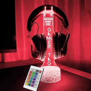 May include: A white LED light stand with a gamer tag design. The stand has a clear acrylic base and a white, textured top. The stand is illuminated with a red light. A pair of black headphones are resting on the stand. A remote control with multiple colored buttons is in the foreground.