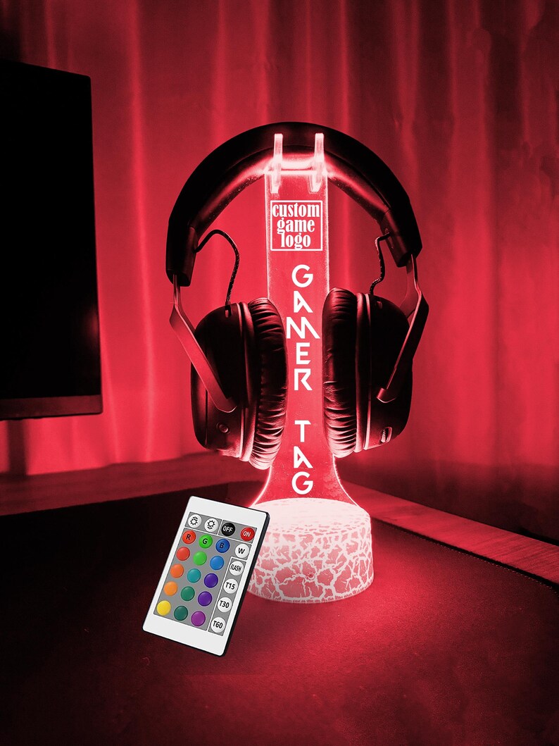 May include: A white LED light stand with a gamer tag design. The stand has a clear acrylic base and a white, textured top. The stand is illuminated with a red light. A pair of black headphones are resting on the stand. A remote control with multiple colored buttons is in the foreground.