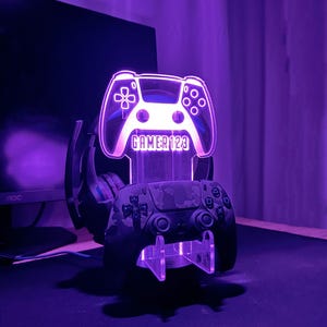 May include: A purple illuminated gaming controller stand with a gamertag "GAMER123" on the front. A black and camouflage gaming controller and a black headset are on the stand.