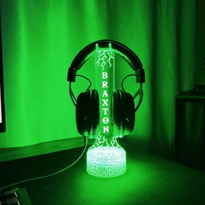 May include: A black headset rests on a custom-made, illuminated headphone stand. The stand is green and features the name "BRAXTON" vertically, with a lightning bolt design. The base of the stand has a textured, glowing pattern.
