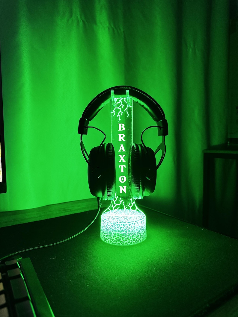 May include: A black headset rests on a custom-made, illuminated headphone stand. The stand is green and features the name "BRAXTON" vertically, with a lightning bolt design. The base of the stand has a textured, glowing pattern.