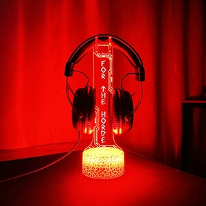 May include: A red illuminated headphone stand with the text "FOR THE HORDE". The stand is holding a pair of black headphones. The base of the stand has a textured, glowing design.