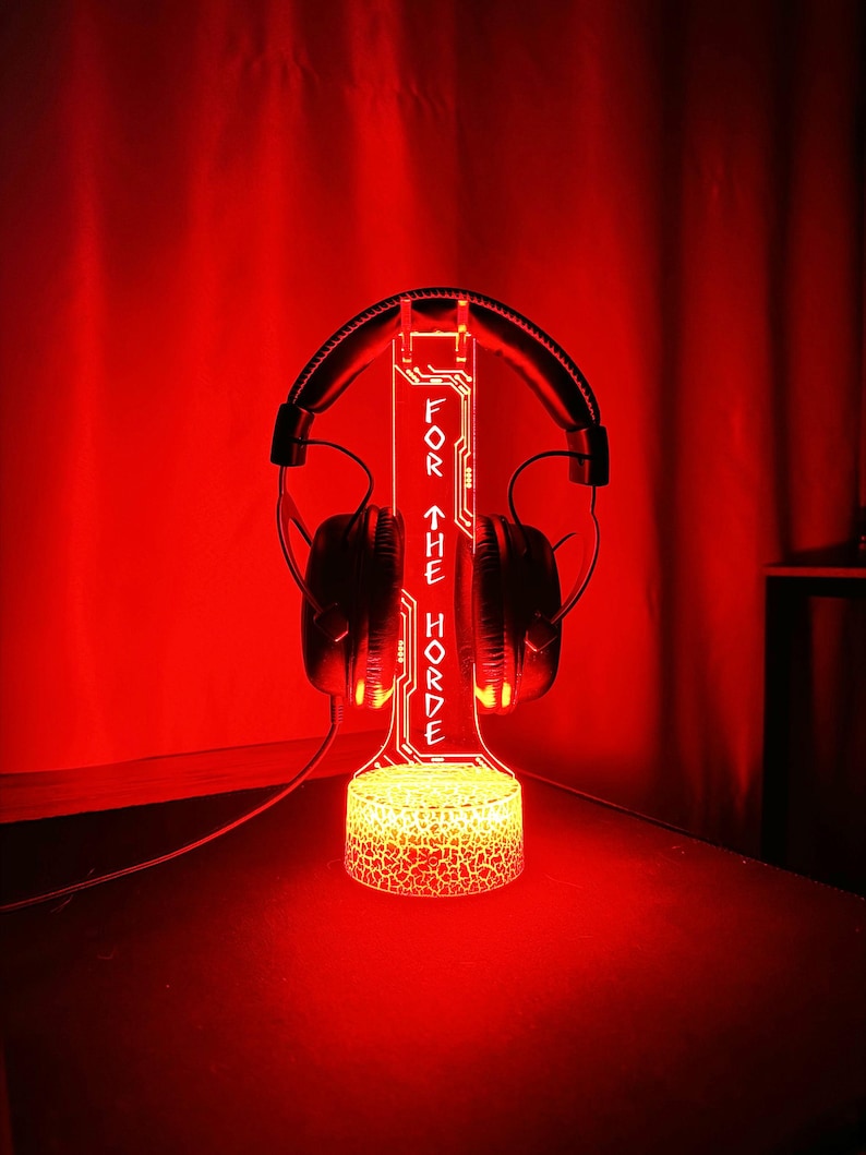 May include: A red illuminated headphone stand with the text "FOR THE HORDE". The stand is holding a pair of black headphones. The base of the stand has a textured, glowing design.