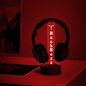 May include: A black and red 3D printed LED lamp with the name "RashNezz" printed on it. The lamp is shaped like a tall cylinder and is sitting on a black base. A pair of black headphones are resting on the top of the lamp.