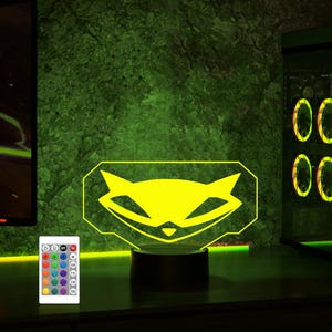 May include: A yellow raccoon logo illuminated on a black base with a remote control for changing the color of the light. The logo is a stylized raccoon face with a mischievous grin.