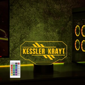 Pu&ograve; includere: Una luce LED gialla con una base nera. La luce presenta un design personalizzato con il testo "Kessler Krayt". La luce &egrave; accesa e illumina l'area circostante.