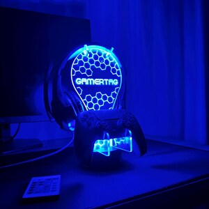 May include: A blue LED light stand with a honeycomb design and the word "GAMERTAG" in white. The stand holds a black video game controller.