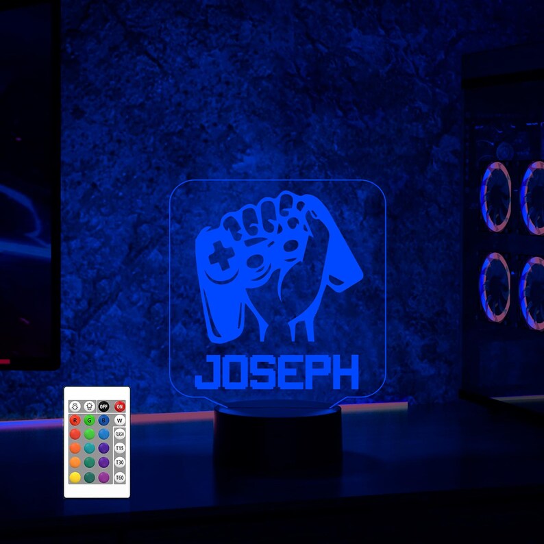 May include: A blue and white personalized LED night light with a gamer hand holding a controller. The name "JOSEPH" is printed below the image. The light is on a black base and has a remote control.