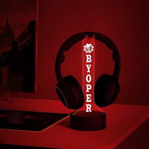 Op de afbeelding: Een rood LED-licht met een wit schedel- en kruisbeenderlogo en het woord "BYOPER" in witte letters. Het licht staat op een zwarte basis en wordt omringd door een zwarte koptelefoon.