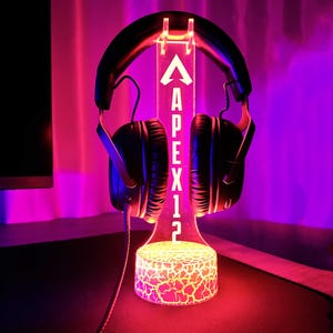 May include: A black gaming headset with a white LED stand that says "Apex" on it. The stand has a cracked glass effect and is lit up in pink and red.