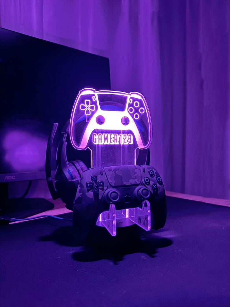 Op de afbeelding: Een paars verlichte gaming controller standaard met de tekst "GAMER123" bovenaan. Een camouflage-patroon controller rust op de standaard, met een headset erachter. De standaard staat op een zwart oppervlak.