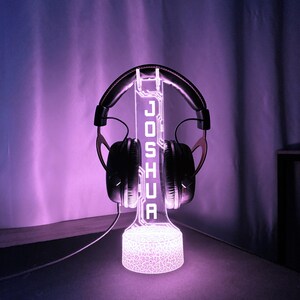 May include: A glowing purple headphone stand with the name "JOSHUA" illuminated in white. The stand holds a pair of black headphones. The base of the stand has a textured pattern, and the background is a gradient of purple.