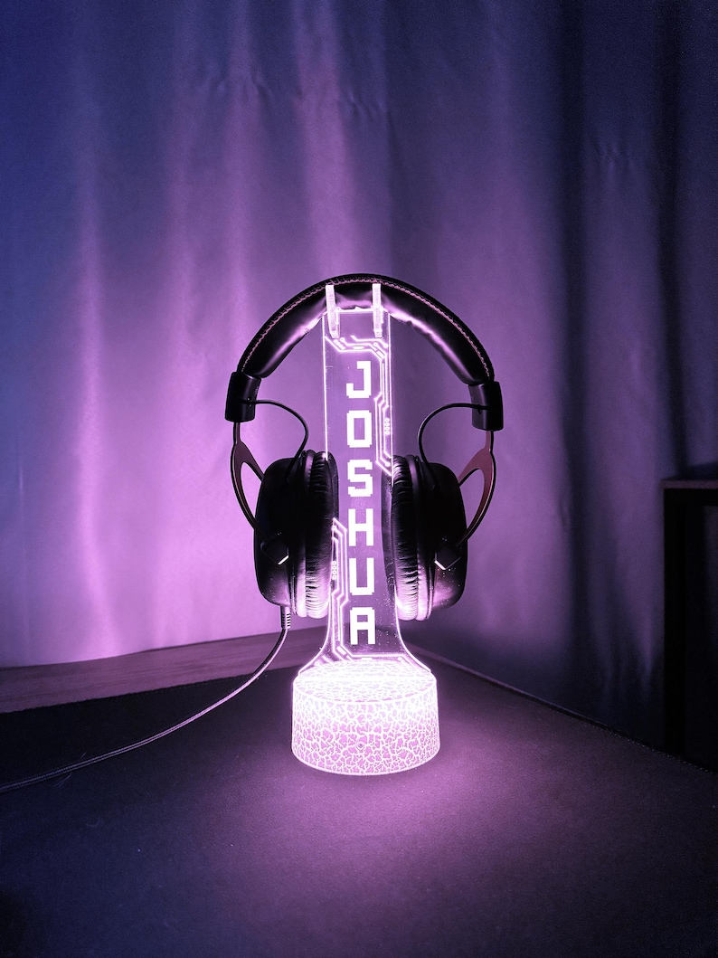 May include: A glowing purple headphone stand with the name "JOSHUA" illuminated in white. The stand holds a pair of black headphones. The base of the stand has a textured pattern, and the background is a gradient of purple.