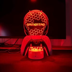 May include: A red LED light with a honeycomb design and the word "GAMERTAG" in yellow letters. The light is mounted on a white stand with two white controllers on either side.