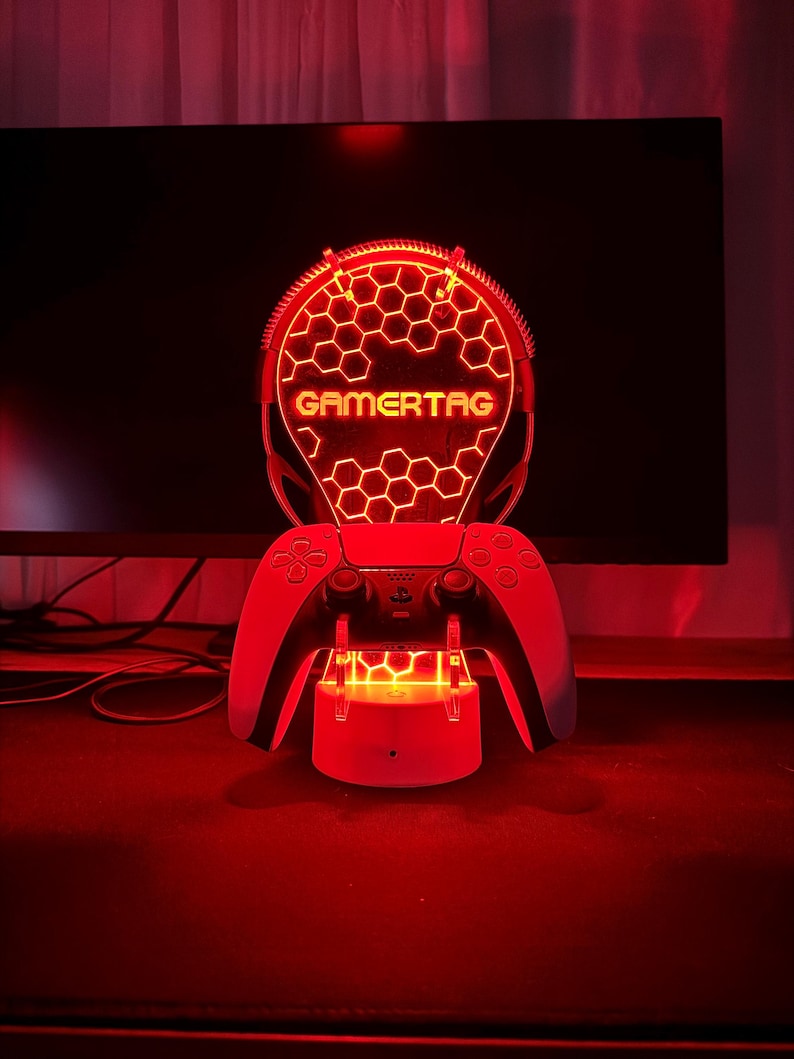 May include: A red LED light with a honeycomb design and the word "GAMERTAG" in yellow letters. The light is mounted on a white stand with two white controllers on either side.