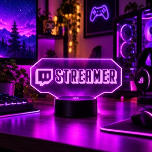 May include: A glowing purple LED sign featuring the Twitch logo and the word STREAMER in bold, block letters. The sign is mounted on a circular black base with a power button, emitting a vibrant neon light across the desk surface.