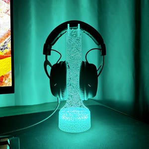 May include: A black gaming headset rests on a glowing blue LED headphone stand. The stand has a detailed topographic design and a circular base. The background is a teal color.