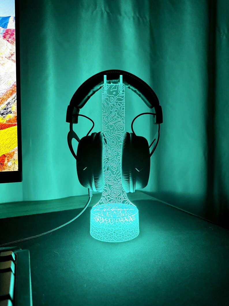 May include: A black gaming headset rests on a glowing blue LED headphone stand. The stand has a detailed topographic design and a circular base. The background is a teal color.