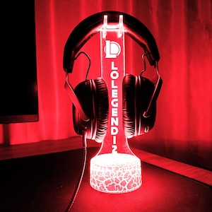 May include: A black gaming headset rests on a glowing red LED stand. The stand features the text "LOLEGENDIN" and a logo. The base of the stand has a cracked pattern design, and the background is a deep red.