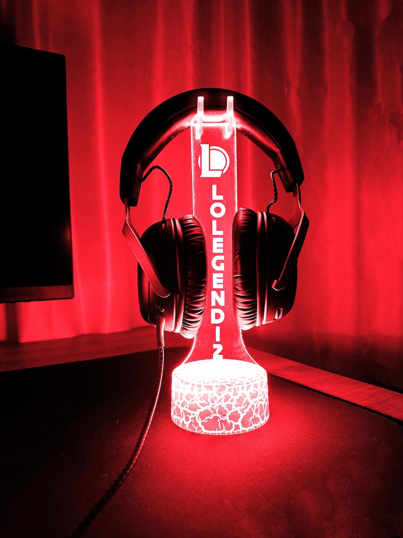 May include: A black gaming headset rests on a glowing red LED stand. The stand features the text "LOLEGENDIN" and a logo. The base of the stand has a cracked pattern design, and the background is a deep red.