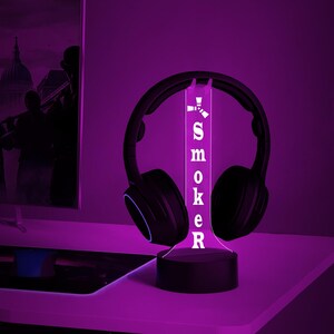 May include: A black and white 3D printed lamp with the word "SMOKER" printed on it. The lamp is lit up in a purple color and has a pair of black headphones resting on it.