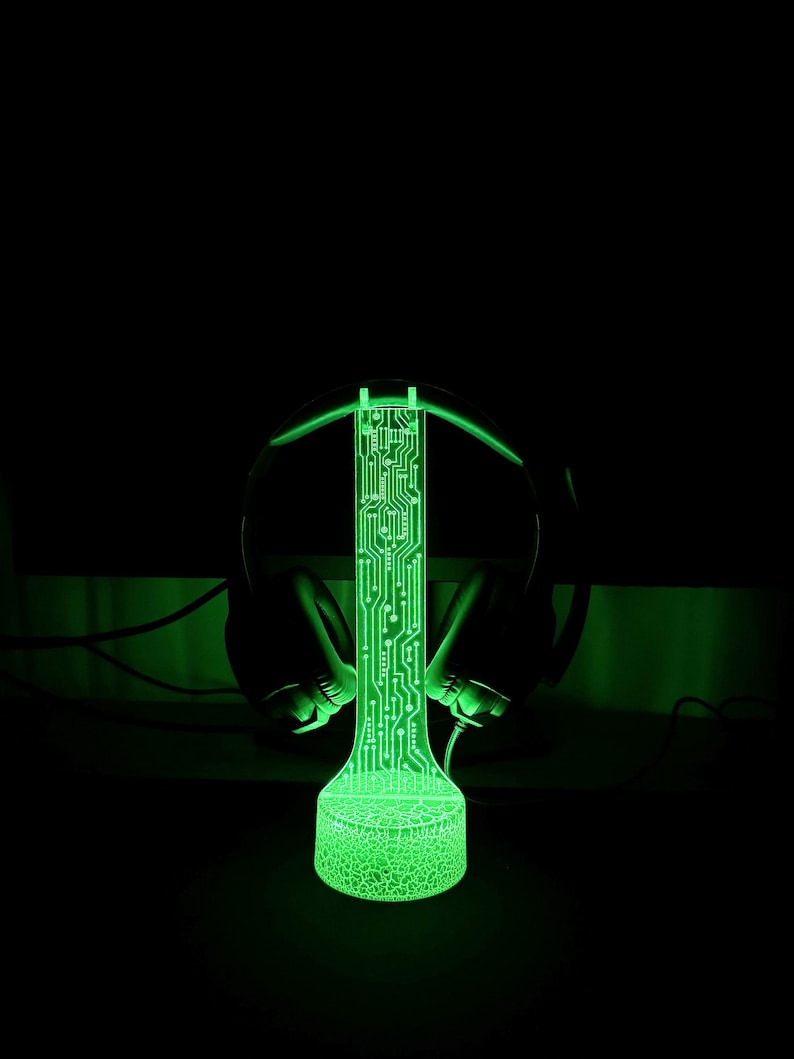 Op de afbeelding: Een groene, gloeiende headsetstandaard met een printplaatontwerp. De standaard is verlicht met groen licht en heeft een ronde basis.