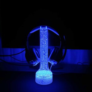 Op de afbeelding: Een blauwe LED-lamp met een printplaatontwerp, die op een standaard staat met een headset erop. De lamp is aan en geeft een blauwe gloed af.