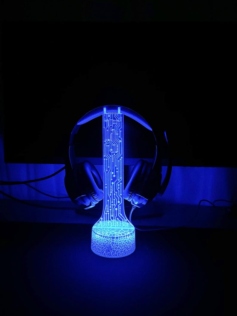 Op de afbeelding: Een blauwe LED-lamp met een printplaatontwerp, die op een standaard staat met een headset erop. De lamp is aan en geeft een blauwe gloed af.