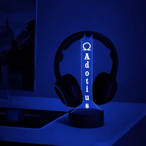 May include: A black headset rests on a custom-made LED stand. The stand is illuminated with a blue glow, displaying the name "Adotius" vertically. The stand's design includes a Greek letter Omega symbol at the top.