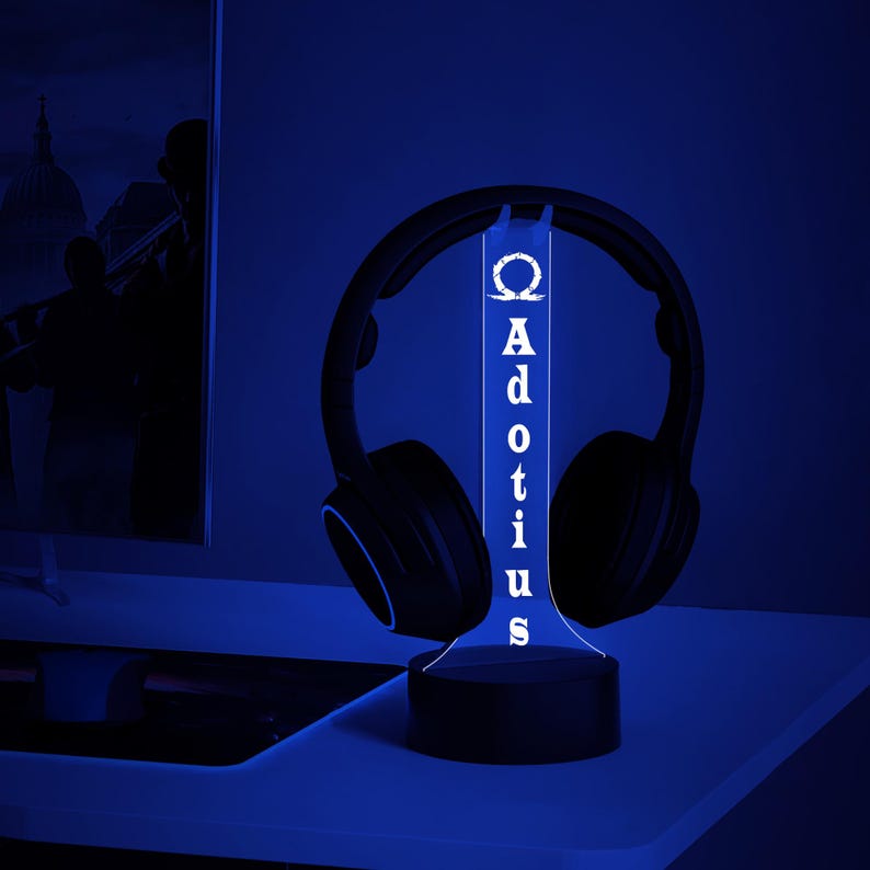 May include: A black headset rests on a custom-made LED stand. The stand is illuminated with a blue glow, displaying the name "Adotius" vertically. The stand's design includes a Greek letter Omega symbol at the top.