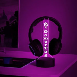 Puede incluir: Una luz LED blanca con una base negra y un panel de acr&iacute;lico transparente que dice "GamerTag" con un s&iacute;mbolo sobre la palabra. La luz est&aacute; encendida e ilumina unos auriculares negros.