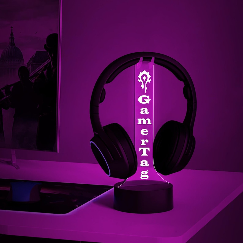 Puede incluir: Una luz LED blanca con una base negra y un panel de acr&iacute;lico transparente que dice "GamerTag" con un s&iacute;mbolo sobre la palabra. La luz est&aacute; encendida e ilumina unos auriculares negros.