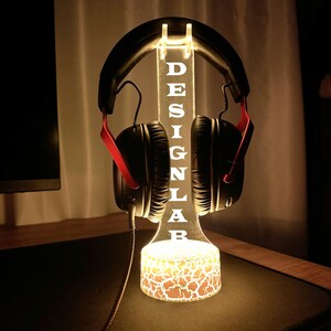 Peut inclure: Un support de casque &eacute;clair&eacute; avec le mot "DESIGN LAB" grav&eacute; dans l'acrylique. Le support est illumin&eacute; d'une lueur chaude, et une paire de casques noirs avec des accents rouges sont affich&eacute;s. La base pr&eacute;sente un motif fissur&eacute;.